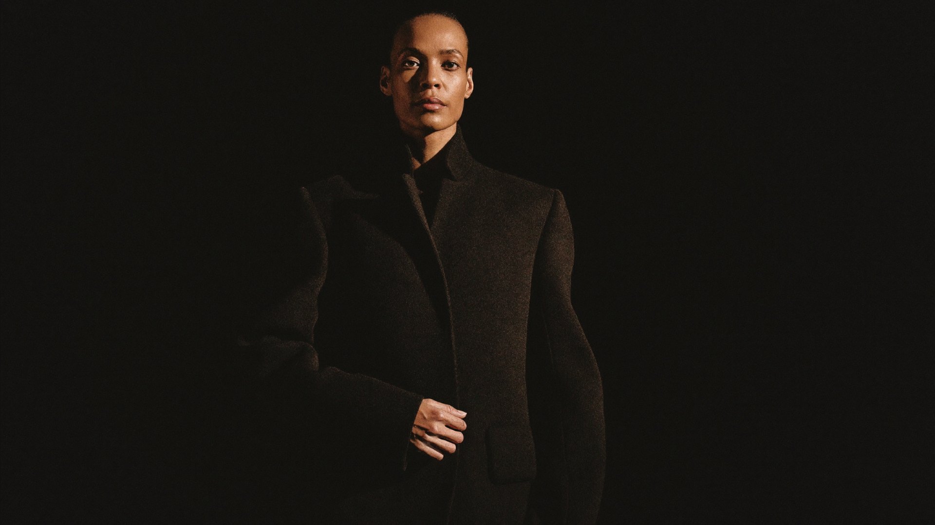 Studio fashion photograph, a model in a sculptural coat under a single hard key light